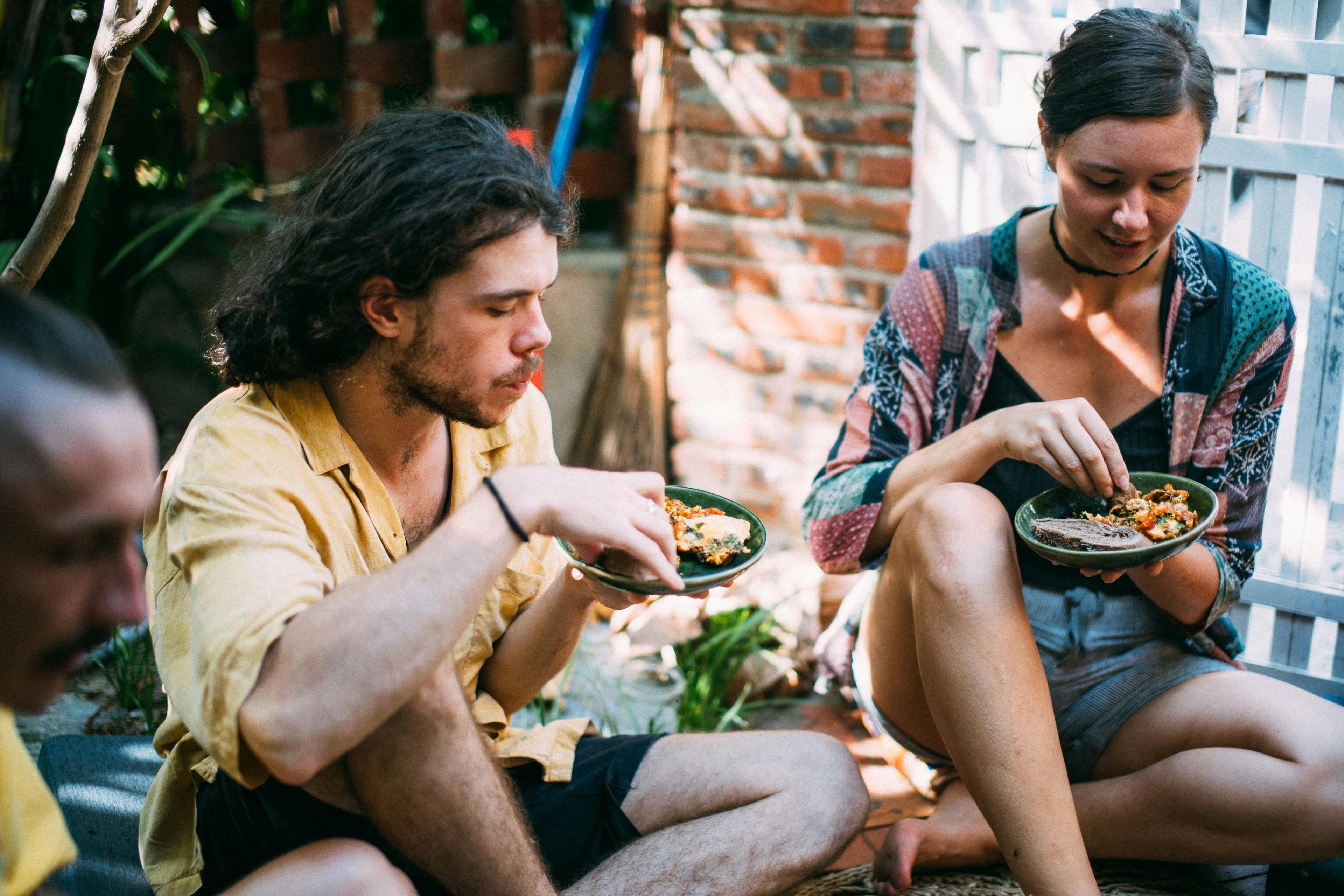 Two young adults sitting cross-legged outdoors sharing a meal — the kind of encounter that closes the distance between strangers.